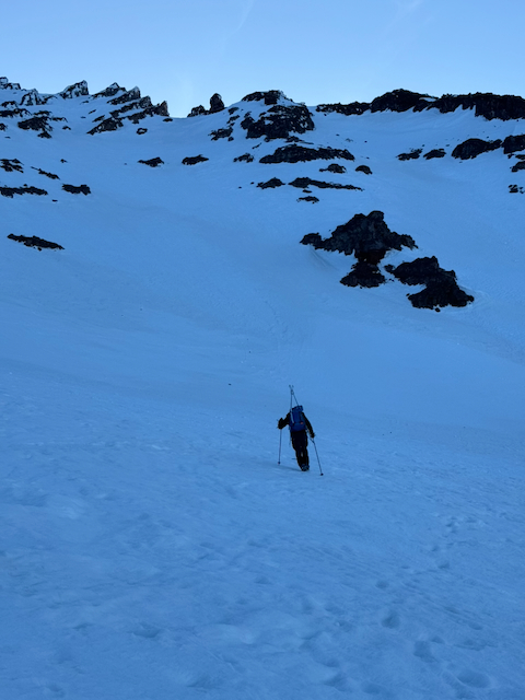 Graham bootpacking up a steep snow gulch in dim pre-dawn blue light, dark volcanic rock formations looming above, skis on his blue pack