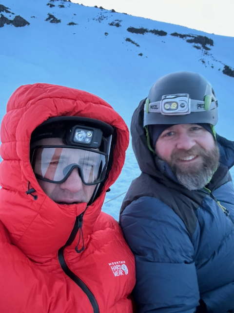 Pre-dawn selfie of Kevin in red puffy with headlamp and Graham in blue puffy with Petzl headlamp on his helmet, both smiling on the snow
