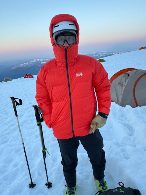 Kevin bundled up in red Mountain Hardwear puffy jacket, helmet, and goggles at camp with tents behind him, sunrise colors on the horizon
