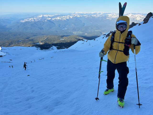 Kevin standing on the steep slope in yellow jacket with skis on pack and crampons, golden sunrise light, sweeping valley and mountain views below