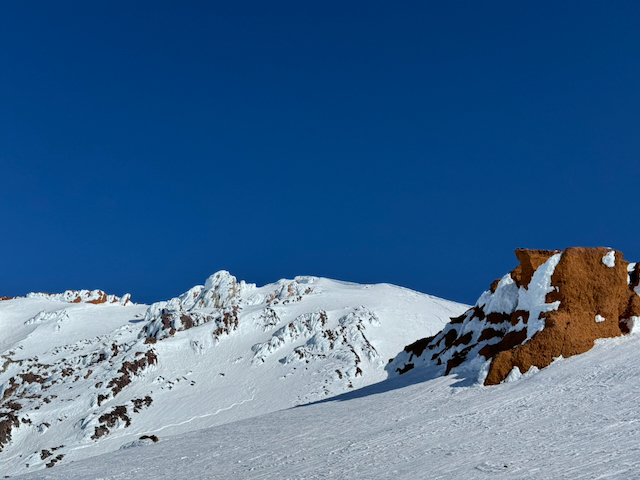 Looking up toward the summit of Shasta — snow-covered peak with dramatic orange volcanic rock towers against deep blue sky