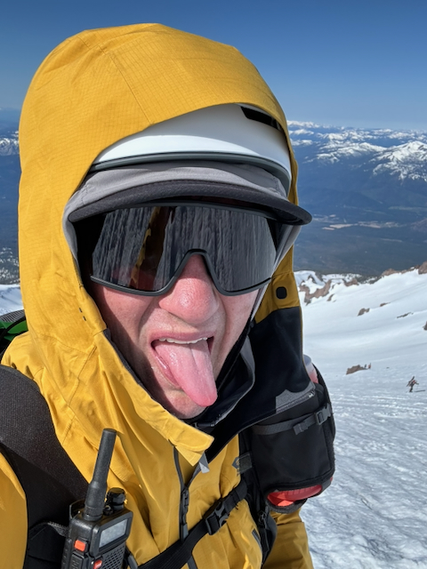 Kevin selfie in yellow jacket sticking his tongue out, white helmet and shield sunglasses, snowy slopes and mountains behind