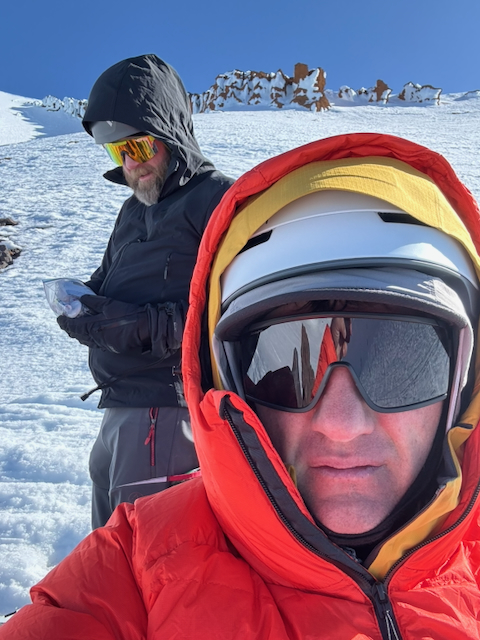 Kevin and Graham selfie near the high point — Kevin smiling in red puffy, Graham in black with orange goggles, volcanic rock formations on the ridge behind