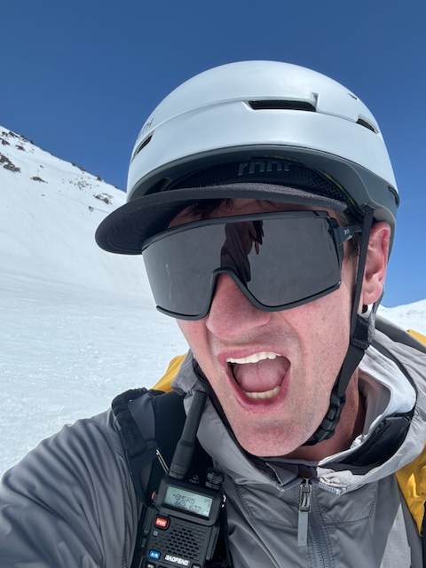 Kevin selfie on the mountain with white helmet, shield sunglasses, and Baofeng radio clipped to his chest, mouth wide open with excitement